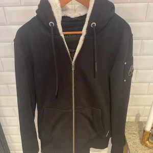 Svart hoodiejacka från Moose Knuckles - Svart hoodiejacka från Moose Knuckles med dragkedja, huva med snörning och vit teddyfoder på insidan. Jackan har två stora fickor framtill och en snygg logga på ärmen. Perfekt för kalla dagar när du vill vara både varm och stilren.