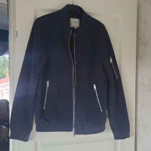 Mörkblå bomberjacka Jack & Jones S - Snygg mörkblå bomberjacka från Jack & Jones med raka linjer och silverfärgade dragkedjor på fickor och ärm. Jackan har klassisk bomberkrage och elastiska muddar vid ärmslut och midja. Perfekt för dig som gillar stilren och enkel design.