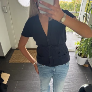 Svart kortärmad blus med knyt - Svart blus med korta ärmar, djup v-ringning och knappar framtill. Toppen har två fickor och ett knytband i midjan som ger en snygg siluett. Perfekt till jeans för en chill look.