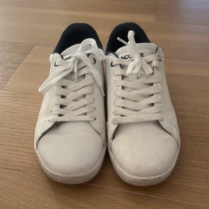 Vita Lacoste sneakers i skinn, strl 37 - Snygga vita sneakers från Lacoste i skinn med klassisk krokodillogga på sidan och mörk detalj vid hälen. Skorna har vita snören, marinblå insida och en röd rand baktill. Perfekta för en clean och sportig look.