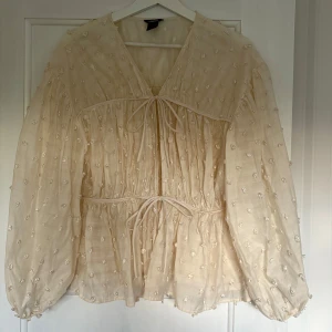 Ljusbeige  blus med broderade prickar - Romantisk beige blus från Lindex med broderade prickar, v-ringning och knytband både vid halsen och midjan. Blusen har långa, luftiga ärmar och voluminös passform. Perfekt för dig som gillar en bohemisk och söt stil.