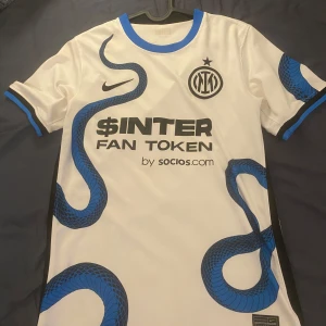 Inter Milan vit bortatröja Nike - Inter Milans vita bortatröja från Nike med blå och svarta ormmönster längs sidorna. Tröjan har klubbmärke, sponsortryck och blå detaljer vid krage och ärmslut. Tillverkad i lätt och ventilerande material, perfekt för match eller träning.