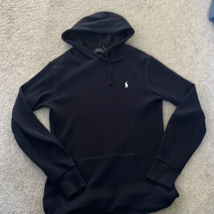 Mörkblå stickad hoodie från Polo Ralph Lauren - Mörkblå stickad hoodie från Polo Ralph Lauren med ribbad struktur och klassisk huva med snörning. Tröjan har lång ärm, känguruficka och den ikoniska vita Polo-loggan broderad på bröstet. Perfekt för chill dagar och snygg streetstyle.