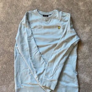 Ljusblå sweatshirt från Lyle & Scott - Ljusblå sweatshirt från Lyle & Scott med klassisk rund hals och broderad gul logga på bröstet. Tröjan har långa ärmar och är tillverkad i mjuk bomull, perfekt för en chill och stilren look. Den är använd ett par gånger men fortfarande klass kvalitet! 