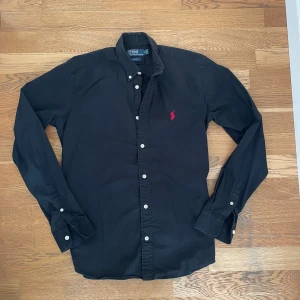 Svart skjorta Polo Ralph Lauren M - Svart skjorta från Polo Ralph Lauren i slim fit med klassisk krage och vita knappar. Broderad röd logga på bröstet. Långärmad modell i bomull som ger en clean och stilren look.
