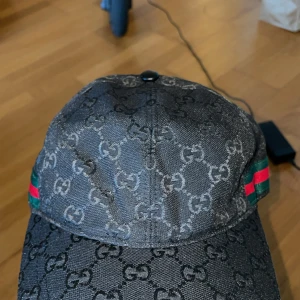 Svart keps från Gucci med GG-mönster - Snygg svart keps från Gucci med klassiskt GG-monogram över hela kepsen och gröna/röda ränder på sidorna. Justerbar rem baktill och tillverkad i polyester. Perfekt accessoar för att lyfta din streetwear-look.