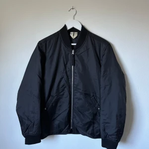 Svart bomberjacka från Arket - Svart bomberjacka från Arket med Thermore-foder för extra värme. Jackan har klassisk bombermodell med ribbade muddar, dragkedja framtill och två snedställda fickor med tryckknapp. Tillverkad i lätt nylonmaterial som ger en clean och stilren look. 3T5