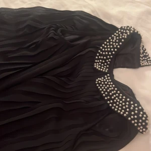 Svart blus  med pärldetaljer - Svart klänning med snygga vågiga veck och en krage dekorerad med pärlor. Klänningen har en elegant känsla och passar perfekt när du vill sticka ut. Materialet känns lätt och flowy.