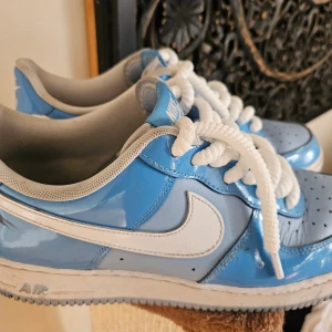 Nike Air Force 1 blå/vita sneaker EU 45 - Nike Air Force 1 sneakers i en snygg blå och vit färgkombination. Skorna har en blank finish, klassisk vit swoosh och tjocka vita snören. Ovandelen är i syntet och skinn, med ventilerande meshfoder och platt sula. Perfekta för dig som gillar streetstyle.