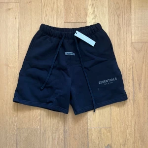 Fear of God Essentials shorts  - Svarta shorts från Fear of God Essentials med elastisk midja och snörning. Mjukt bomullsmaterial, Nice passform och diskret logga framtill. Bakficka och klassisk streetwear-vibe. Perfekta för chill dagar eller när du vill hålla det stilrent och bekvämt. Skriv om ni har frågor eller vill diskutera pris