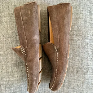 Bruna mocka loafers från GANT - Snygga loafers från GANT i brun mocka med kontrasterande vit söm runt tå och häl. Skorna har en klassisk rund tå och platt sula med GANT-logga på baksidan. Insidan är i ljust skinn och de har en stilren, tidlös design som passar till jeans eller chinos.