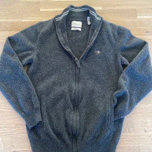 Grå kofta i ull från GANT - Mysig grå kofta från GANT i superfin lammull. Dragkedja framtill, ribbad krage med vita ränder och broderad logga på bröstet. Perfekt för kyliga dagar och enkel att matcha med det mesta.