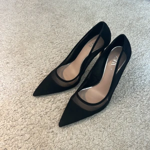 Svarta pumps med spetsig tå från Zara - Snygga svarta pumps från Zara i storlek 38. Skorna har spetsig tå, hög klack och en cool meshdetalj längs sidorna. Tillverkade i en mix av syntet och mocka för en trendig look. Perfekta för dig som vill sticka ut med stilrena detaljer. Ca 9 cm klack. Endast använda 2 gånger 