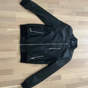 Svart skinnjacka från AC, Made in Italy - Svart skinnjacka från AC, Made in Italy, med flera snygga dragkedjor framtill och klassisk bomberinspirerad passform. Jackan har ribbade muddar vid ärmslut och midja samt slät insida med diskret 'Made in Italy'-tryck.