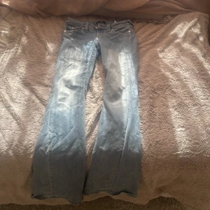 True Religion Joey Low Rise Flare Jeans - Säljer ett par ljusblå True Religion Joey jeans med låg midja och utsvängda ben. Klassiska detaljer med stora bakfickor, dekorativa sömmar och silverfärgade knappar. Jeansen är i denim och har en snygg tvättad look. Dom har hål på insidan men så var de när jag fick som men du syns inte när man har på sig byxorna. De ser lite stora ut på bilden men dom e inte så stora💕