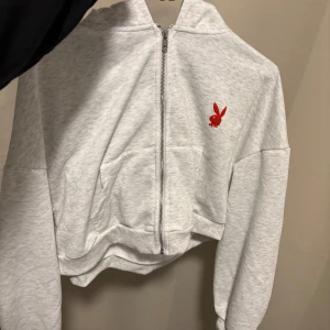 Grå croppad hoodie  - Säljer en ljusgrå croppad hoodie från Playboy med röd logga framtill och stor röd Playboy-text på ryggen. Tröjan har dragkedja, huva och långa ärmar. Perfekt för dig som gillar streetstyle och vill sticka ut.