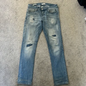 Blå slitna jeans Jack & Jones Glenn - Sällsynta Snygga ljusblå jeans från Jack & Jones, modell slim Glenn. Jeansen har slitna detaljer och hål på benen för en trendig look. Klassisk femficksmodell med knappgylf och smal passform. Perfekta för dig som gillar en avslappnad streetstil.