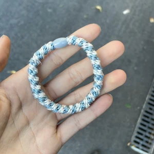Blåvitt flätat armband från Kknekki - Säljer ett snyggt flätat armband från Kknekki i blå och vita toner. Armbandet har en rund form och är gjort av mjukt syntetmaterial med ett litet blått märke. Perfekt att bära varje dag för en chill vibe.