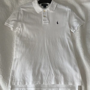 Vit pikétröja från Ralph Lauren L - Klassisk vit pikétröja från Ralph Lauren med broderad marinblå logga på bröstet. Tröjan har korta ärmar, krage och två knappar framtill. Tillverkad i mjuk bomull som andas och passar perfekt till en clean och stilren look.