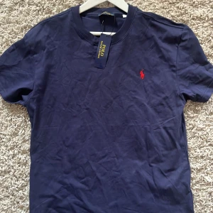 Mörkblå t-shirt från Polo Ralph Lauren - Snygg mörkblå t-shirt från Polo Ralph Lauren med klassisk röd logobrodyr på bröstet. T-shirten har rund hals och korta ärmar, tillverkad i mjuk bomull som känns skön mot huden. Perfekt för dig som gillar stilrena och tidlösa plagg.