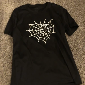Svart t-shirt med spindelnätsprint - Svart t-shirt med vit spindelnätsdesign på bröstet där mitten av nätet är format som ett hjärta. Klassisk rund hals och korta ärmar. Perfekt för dig som gillar edgy och unik stil.