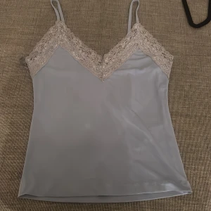 Ljusblå linnetopp med spets Gina Tricot - Supersöt ljusblå linnetopp från Gina Tricot med tunna axelband och v-ringning dekorerad med spets. Toppen har en slät, mjuk yta och är perfekt för lager-på-lager eller sommarens alla outfits.