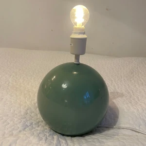 Grön bordslampa med rund fot - Modern bordslampa med rund, grön/blå fot i blank finish. Lampan har vit sockel och tygklädd strömkabel. Fungerar och är i gott skick, inga synliga repor eller skador. Passar utmärkt som dekorativ belysning i hemmet. QC-märkt på undersidan.