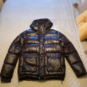 Svart pufferjacka från Urban Pioneers - Snygg svart pufferjacka från Urban Pioneers i storlek M. Jackan har glansig finish, huva, stora fickor med knappar och dragkedja framtill. Väldigt lätt använd, säljer då den inte används. Tre stycken knappar som har lossnat från luvfästet, kan säkert sys för den som är kunnig. Luvan är fullt användbar ändå och den kan enkelt tas bort.