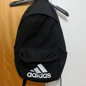 Svart Adidas ryggsäck med logga - Säljer en svart ryggsäck från Adidas med stor vit logga framtill. Väskan har ett stort huvudfack, en framficka med dragkedja och två sidofickor. Tillverkad i slitstark polyester, perfekt för skola eller träning. Enkel och stilren design som funkar till det mesta. lite sliten och har ett litet hål som lätt går att fixa