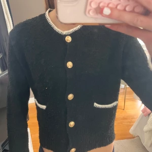 Svart kofta med guldknappar från H&M - Svart kofta från H&M Divided med guldiga dekorativa knappar och vita detaljer vid krage och fickor. Koftan är stickad och har lång ärm, perfekt för lager-på-lager. Klassisk och snygg modell som lyfter din outfit.