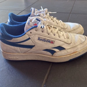 Reebok skor sneaker blå - Klassiska Reebok sneakers i vitt skinn med blå detaljer och logga på sidan och hälen. Skorna har snörning, rund tå och platt sula. Insidan är fodrad med blå textil för extra komfort. Perfekta för dig som gillar retrostil och sportig vibe.