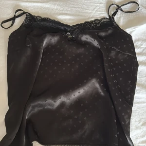 svart satin pyjamas från newyorker! - Supersöt svart linnetopp i glansigt satinliknande material med små hjärtan som mönster. Toppen har tunna axelband, spetskant upptill och en liten rosett framtill. Perfekt för dig som gillar romantiska detaljer och en mjuk känsla mot huden. Shortsen är även matchande fast utan hjärtan!