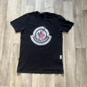 Svart Moncler t-shirt med stor logga - Svart t-shirt från Moncler Grenoble med stor broderad logga i vitt, blått och rött på bröstet samt mindre logga på ärmen. Klassisk passform och rund halsringning. Tillverkad i mjuk bomull för en skön känsla.