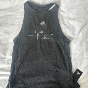 Mörkgrå/svart Adidas träningsstopp - Snygg mörkgrå linnetopp från Adidas med stort logotryck framtill. Toppen har ärmlös design, rundad halsringning och liten slits på båda sidorna. Perfekt för träning eller chill dagar. Mjukt och stretchigt material