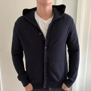 Mörkblå stickad kofta från Less Worries  - Mörkblå stickad kofta med huva och knäppning framtill. Koftan har två fickor och långa ärmar. Enkel och stilren design som passar till det mesta. Nästan aldrig använd, nypris 1799.