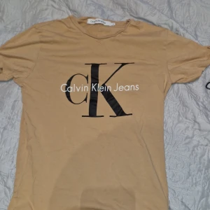 Beige Calvin Klein Jeans t-shirt XS - Snygg beige t-shirt från Calvin Klein Jeans i bomull med stor svart CK-logga och vit text framtill. Klassisk rund hals och korta ärmar. Perfekt för dig som gillar stilrena och trendiga plagg med tydlig logga.