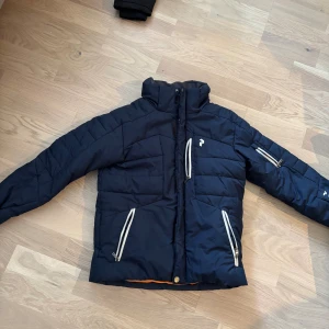 Mörkblå Peak Performance pufferjacka 130 - Snygg mörkblå pufferjacka från Peak Performance med orange foder. Jackan har flera dragkedjeförsedda fickor och hög krage. Perfekt för kalla dagar och har en sportig vibe. Juniorstorlek 130.