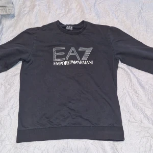 Svart EA7 Emporio Armani sweatshirt - Svart sweatshirt från EA7 Emporio Armani med stor vit logga framtill. Klassisk rund halsringning och långa ärmar. Mjuk och skön bomullskänsla, perfekt för en stilren och avslappnad look.
