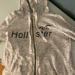 Grå hoodie med dragkedja från Hollister - Mysig ljusgrå hoodie från Hollister med broderad logga och text på bröstet samt 'Los Angeles' på ärmen. Tröjan har huva med snörning, dragkedja framtill och kängurufickor. Perfekt för chill dagar och streetstyle.