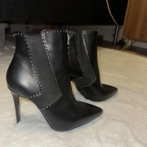 Svarta stövletter med nitar från Derimod - Snygga svarta stövletter från Derimod i skinnimitation med spetsig tå och hög klack. Skorna har dekorativa små nitar längs kanterna och elastiska paneler på sidorna för enkel påtagning. Perfekta för dig som vill ha en edgy och trendig look.