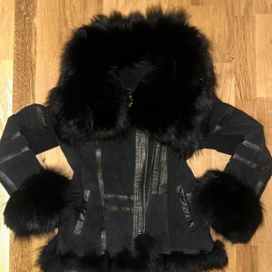Svart pälsjacka med stora detaljer - Säljer en svart pälsjacka med stora fluffiga pälskanter vid krage, ärmslut och nederkant. Jackan har snygga detaljer i skinn och mocka, samt dragkedja framtill. Perfekt för dig som vill sticka ut och hålla dig varm under kalla dagar.