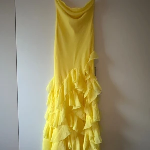Gul långklänning med volanger - Superfin gul långklänning med tunna axelband och snygg draperad urringning. Klänningen har flera lager volanger nedtill som ger en riktigt flowy känsla. Materialet är lätt och luftigt, perfekt för varma dagar. Knytband i ryggen för extra detalj.