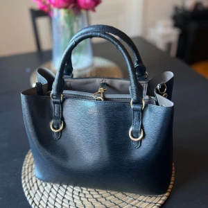 Svart handväska från Lauren Ralph Lauren - Snygg svart handväska i strukturerat skinn från Lauren Ralph Lauren. Väskan har guldfärgade metalldetaljer, dubbla handtag och avtagbar axelrem. Insidan är ljusbeige och har dragkedja.  Obs: Handtaget har ett litet slitage (syns på bild), men påverkar inte användningen. #ralphlauren