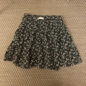 Blommig svart kortkjol från Lager 157 - Supersöt svart kortkjol från Lager 157 med vitt blommigt mönster. Kjolen har volangdetaljer och är tillverkad i mjuk polyester med lite stretch för skön passform. Perfekt för dig som gillar en lekfull och trendig stil.