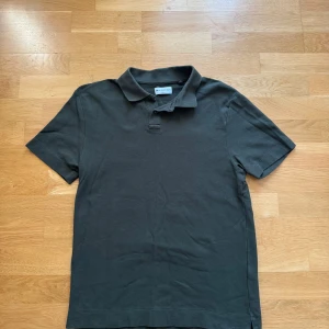 Mörkgrå pikétröja från These Glory Days - Snygg mörkgrön pikétröja från These Glory Days i klassisk modell med krage och knappar. Tillverkad i mjuk bomull och har korta ärmar. Perfekt för en clean och stilren look.