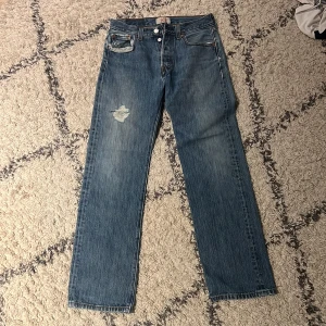Levi's 501 blå jeans straight fit - Klassiska Levi's 501 jeans i blå denim med raka ben och knäppning med knappar. Jeansen har en sliten detalj på vänster lår och femficksdesign. Perfekt för dig som gillar en tidlös och avslappnad look. Dock trasig högerficla men den är lätt att sy ihop!!