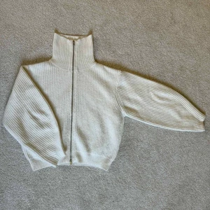 Beige ribbad kofta från BikBok - Supermysig beige ribbad kofta från BikBok med hög krage och hel dragkedja framtill. Tröjan är stickad i ett mjukt material och har långa ärmar med breda muddar. Perfekt för lager-på-lager under kyliga dagar.