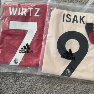 Adidas fotbollströjor Wirtz & Isak - Två snygga fotbollströjor från Adidas med tryck: en röd Wirtz #7 och en beige Isak #9. Båda har stora siffror och namn på ryggen, samt Premier League-logga. Perfekt för dig som vill sticka ut på planen eller visa support för dina favoritspelare.