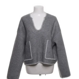 Grå kofta från Zara med vita detaljer - Grå kofta från Zara i medium med vita dekorativa sömmar längs kanterna. Tröjan har två stora fickor framtill, v-ringad hals och långärmade, loose passform. Materialet är mjukt och känns ulligt, perfekt för kyliga dagar.