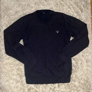 Mörkblå stickad tröja från GANT - Säljer en klassisk mörkblå stickad tröja från GANT med rund halsringning och diskret logga broderad på bröstet. Nypris: 1100